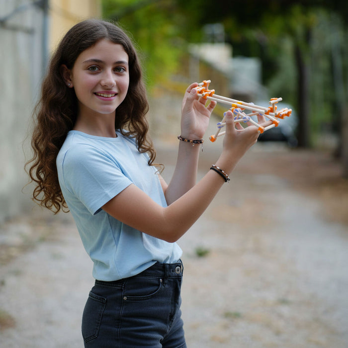 A young woman with long brown curly hair stands outdoors holding a small wooden structure from beaver with orange and white details. She is wearing a light blue t-shirt and dark jeans, with a blurred background of trees and a path.