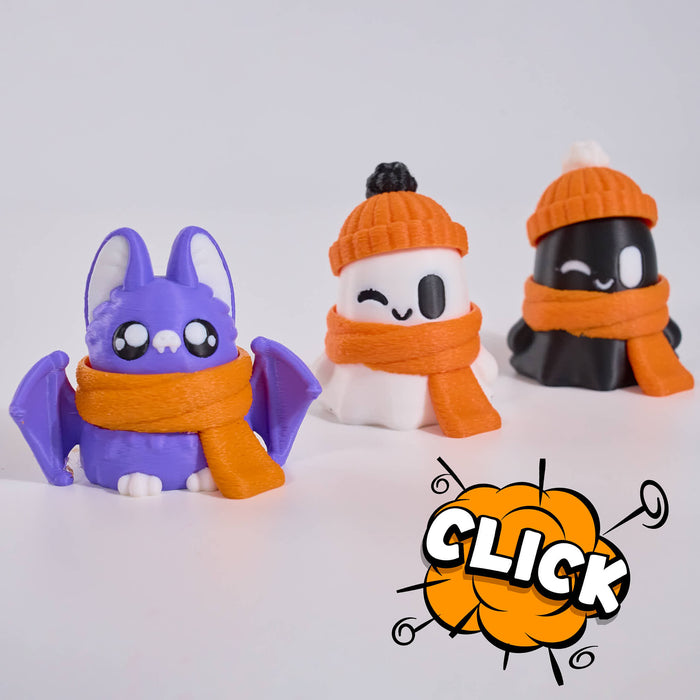 Three cute, stylized character figurines wearing orange knit beanies and scarves, arranged on a clean white studio background. The left figure is purple with bat wings and large white eyes; the center figure is white with a winking eye and black nose; the right figure is black with a winking eye.