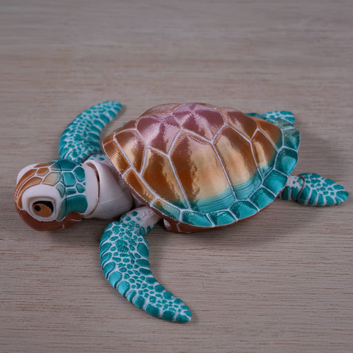 A small decorative sea turtle figurine with a metallic bronze and gold shell, turquoise and teal flippers, and a white head, photographed on a light wood surface.