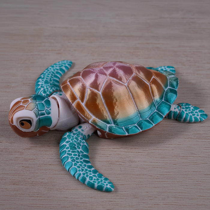 A small decorative sea turtle figurine with a metallic bronze and gold shell, turquoise and teal flippers, and a white head, photographed on a light wood surface.
