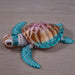 A small decorative sea turtle figurine with a metallic bronze and gold shell, turquoise and teal flippers, and a white head, photographed on a light wood surface.