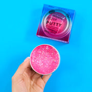 Pink glittery putty in a container held by a hand against a blue background