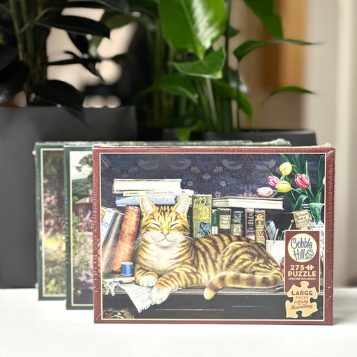 Cobble Hill puzzle box with a cat and books illustration on a surface with plants in the background