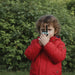 A young child wearing a bright red puffer jacket stands outdoors in front of a green hedge, holding a small walkie-talkie to their face from kidywolf