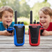 Two children holding toy walkie-talkies are smiling in the background behind two larger walkie-talkies standing on a wooden table. The walkie-talkies are blue and red with black antennas and black front panels.