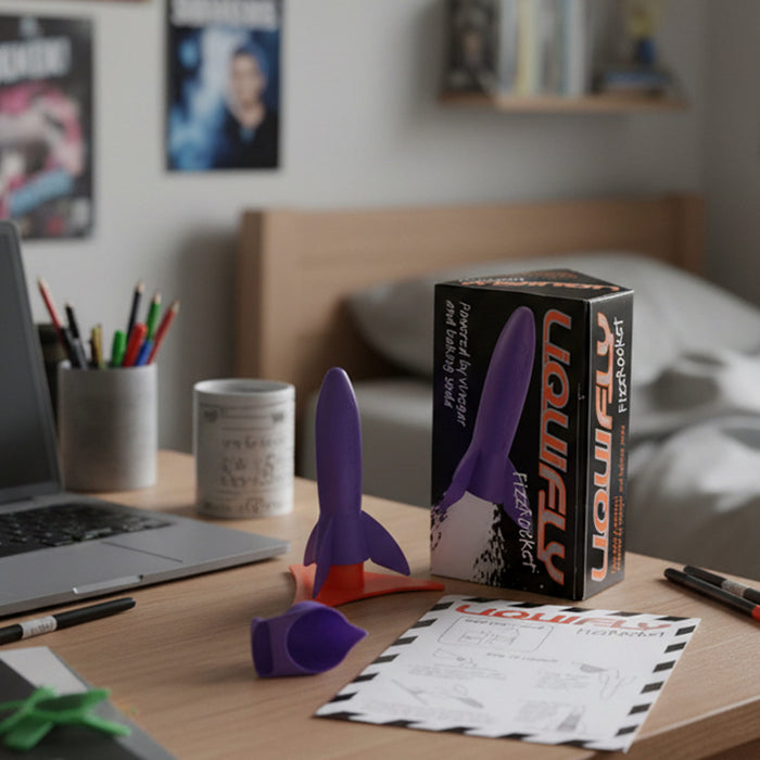 A purple heebie Jeebies  liquifly rocket-shaped with an instructions  displayed on a wooden desk next to its black and orange retail box, with a laptop, mug, and bedroom background visible.