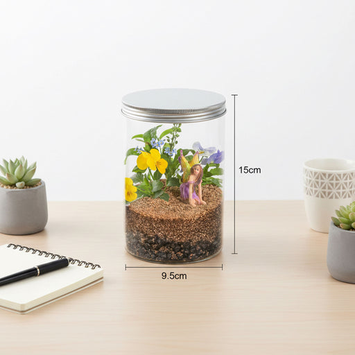 A clear glass jar terrarium with a silver metal lid sits on a light wood desk. The jar contains brown soil, green leaves, yellow and purple flowers, and a small fairy figurine.
