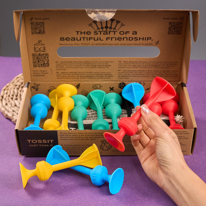 Cardboard packaging of TOSSIT product with colorful elements and text. Colorful dog toys with a hand holding a red toy, displayed in front of a Tossit packaging box.