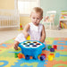 Quercetti Daisy Shape Sorter baby toy with toddler sits on a colorful foam play mat in a bright nursery, playing with a blue flower-shaped sorting toy and colorful geometric blocks.