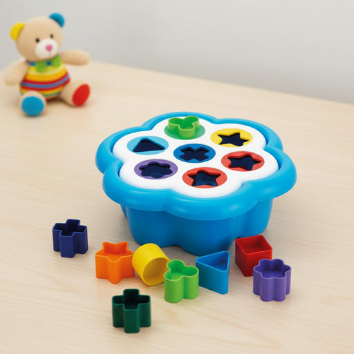 Quercetti Daisy Shape Sorter baby toy in  colorful plastic shape-sorting toy set on a light wood surface, with a blue flower-shaped tray holding multicolored geometric pieces and additional loose pieces arranged around it. A small teddy bear toy sits blurred in the background.