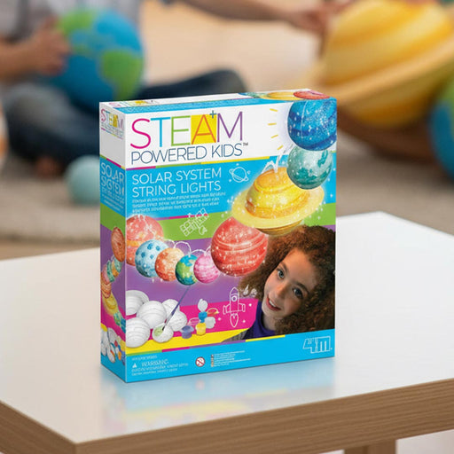 STEAM Powered Kids Solar System String Lights toy box displayed on a white table in a blurred indoor setting with children playing in the background.
