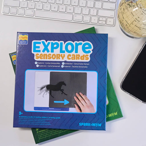 A boxed activity product labeled 'EXPLORE Sensory Cards' lies on a white desk, partially covering green activity sheets. A keyboard, a globe, and a smartphone are visible around it.