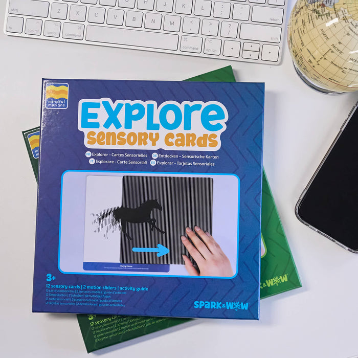 A boxed activity product labeled 'EXPLORE Sensory Cards' lies on a white desk, partially covering green activity sheets. A keyboard, a globe, and a smartphone are visible around it.