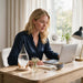 A woman with blonde hair sits at a wooden desk in a bright room, looking at a laptop.  on her side the heebie jeebies teardrop