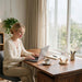 A woman in a cream cardigan and beige pants sits at a wooden desk using a laptop in a bright home office setting with a window, curtains, and a Fitzroy's Storm Glass Teardrop Shape
