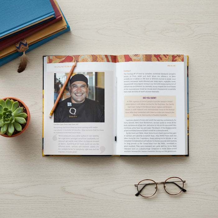 The Little Red, Yellow, Black Book: An introduction to Indigenous Australia, 4th Edition  laid flat on a light wood surface, showing printed text and a portrait photo of a smiling man. A pencil rests on the pages, with stacked books, a small succulent, and eyeglasses around it.