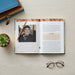 The Little Red, Yellow, Black Book: An introduction to Indigenous Australia, 4th Edition  laid flat on a light wood surface, showing printed text and a portrait photo of a smiling man. A pencil rests on the pages, with stacked books, a small succulent, and eyeglasses around it.