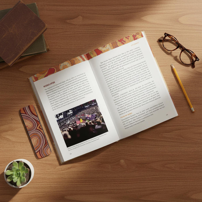 The Little Red, Yellow, Black Book: An introduction to Indigenous Australia, 4th Edition laid on a wooden desk with printed text and a small embedded photo on the left page. The book has a decorative multicolor patterned cover and matching patterned interior accents.