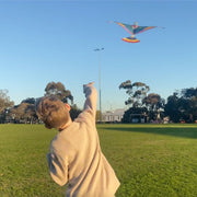 Child flying a colorful kite in an open park with trees and buildings in the background.
