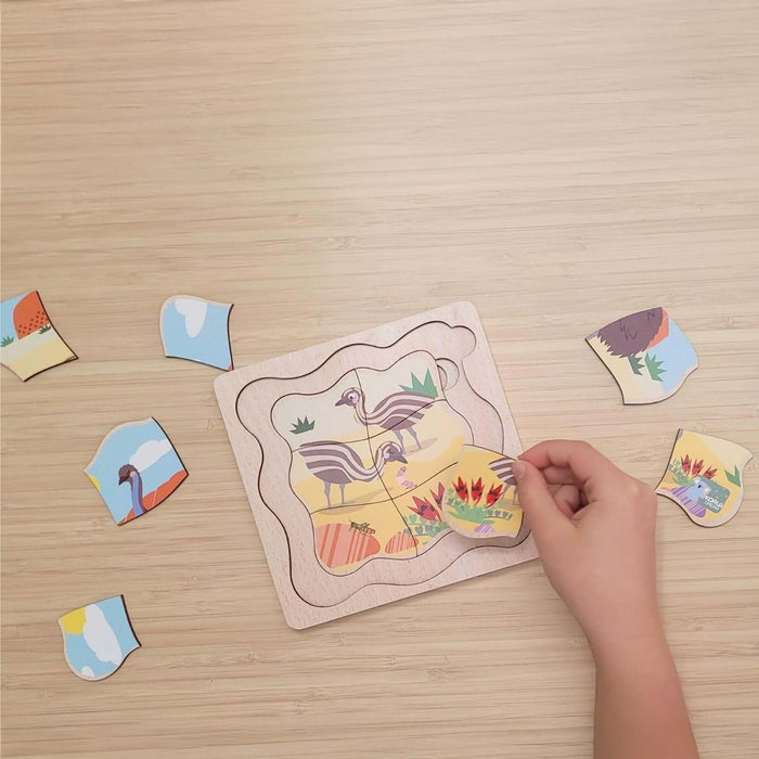 A wooden puzzle board with illustrated animal and landscape pieces being assembled on a light wood tabletop by a person's hand. Several loose puzzle pieces are scattered around the board.