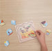 A wooden puzzle board with illustrated animal and landscape pieces being assembled on a light wood tabletop by a person's hand. Several loose puzzle pieces are scattered around the board.