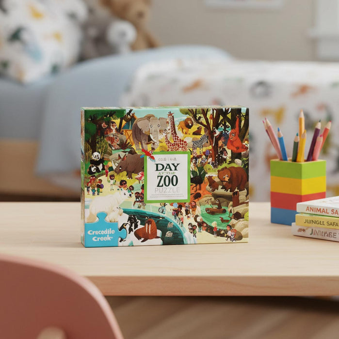A jigsaw puzzle box titled 'The Day Zoo' by Crocodile Creek sits on a light wood desk in a child's room, with colorful pencils in a block cup and books in the background.