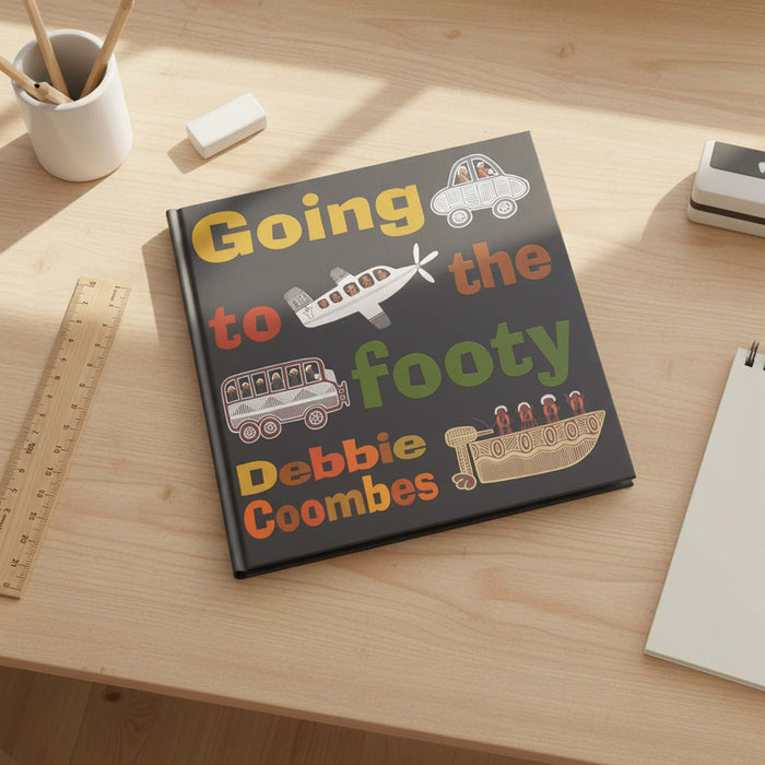 A hardcover children's book titled 'Going to the footy' by Debbie Coombes lies open on a light wood desk. The cover is dark gray with colorful illustrations of a car, airplane, bus, and boat, plus large multicolor text.