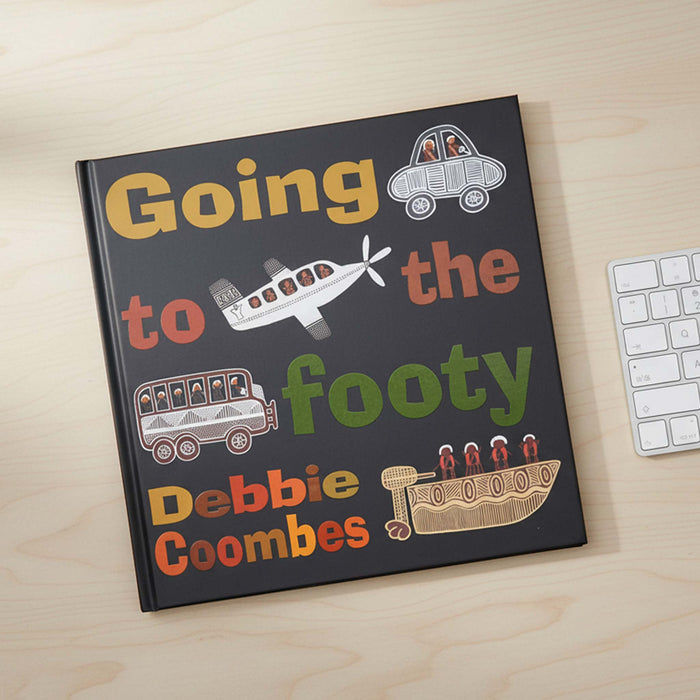 A hardcover book titled 'Going to the footy' by Debbie Coombes lies open on a light wood desk next to a white keyboard. The cover is dark charcoal with colorful hand-drawn illustrations of a car, airplane, bus, and boat, plus multicolored text.