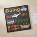 A hardcover book titled 'Going to the footy' by Debbie Coombes lies open on a light wood desk next to a white keyboard. The cover is dark charcoal with colorful hand-drawn illustrations of a car, airplane, bus, and boat, plus multicolored text.