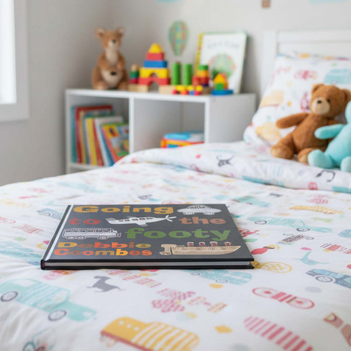 A children's board book titled 'Going to the Footy Cribbits' placed on a colorful patterned duvet in a child's bedroom, with toys and a teddy bear visible in the softly blurred background.