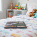 A children's board book titled 'Going to the Footy Cribbits' placed on a colorful patterned duvet in a child's bedroom, with toys and a teddy bear visible in the softly blurred background.