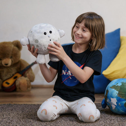 A young child sits on a carpeted floor in a cozy room, holding a plush moon toy. The child is wearing a black t-shirt with a NASA logo and white leggings with rainbow patterns.