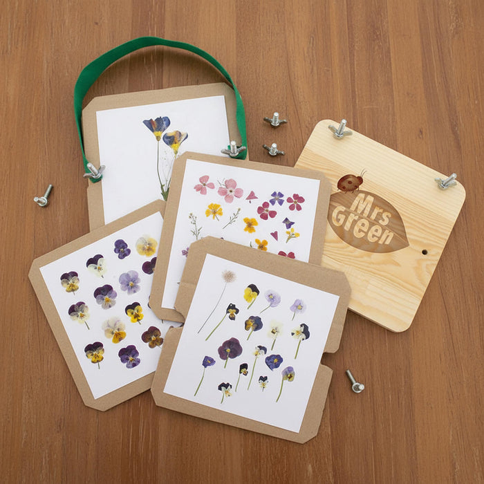 A set of pressed flower cards and a wooden hanging board on a wood tabletop. The cards have white paper inserts with pressed flowers on brown kraft backing, and one wooden board has a green ribbon handle and the words 'Mrs Green' printed on it.