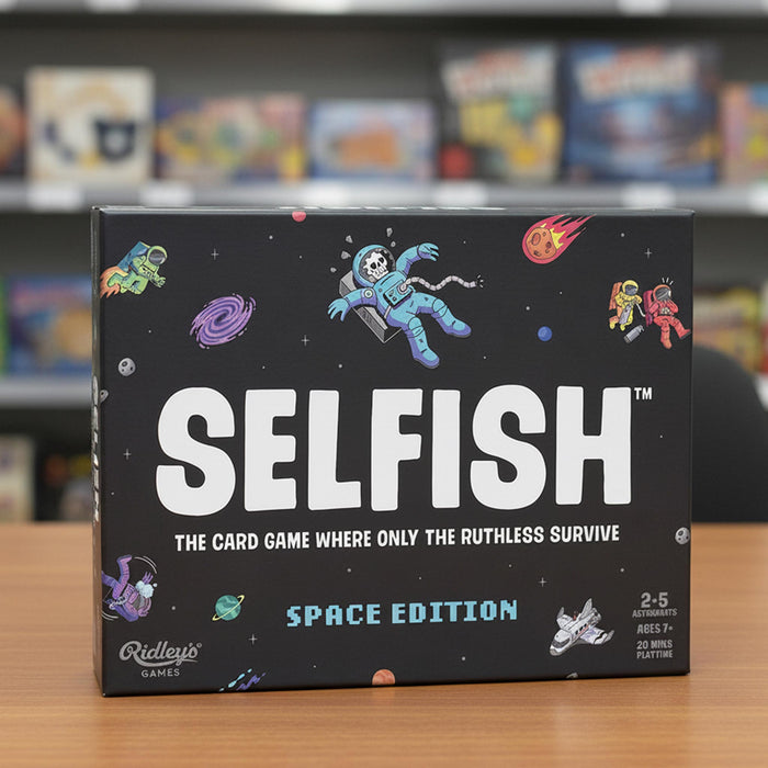 The board game box for 'Selfish: Space Edition' by Ridley's Games, featuring a black background decorated with colorful cartoon astronauts, planets, and stars. The box is displayed on a wooden table with a blurred store shelf in the background.