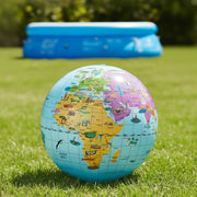 A colorful world map globe resting on green grass, with a blue inflatable pool blurred in the background outdoors.