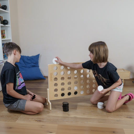 Two children playing with a large wooden connect-four game on a wooden floor.